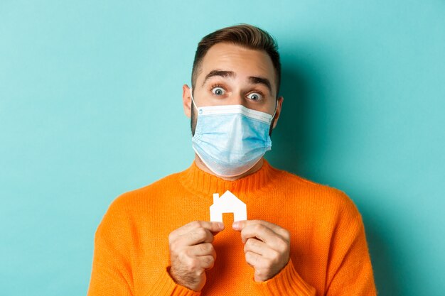 Real estate and coronavirus pandemic concept. Close-up of adult man in medical mask holding small house