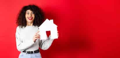 Free photo real estate excited smiling woman showing paper house cutout and looking amazed standing on red back