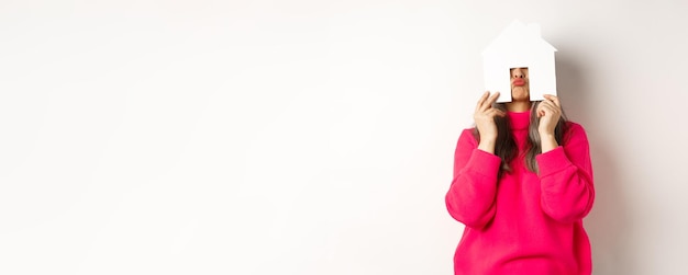 Real estate funny asian middleaged woman hiding face behind paper house model and showing puckered l