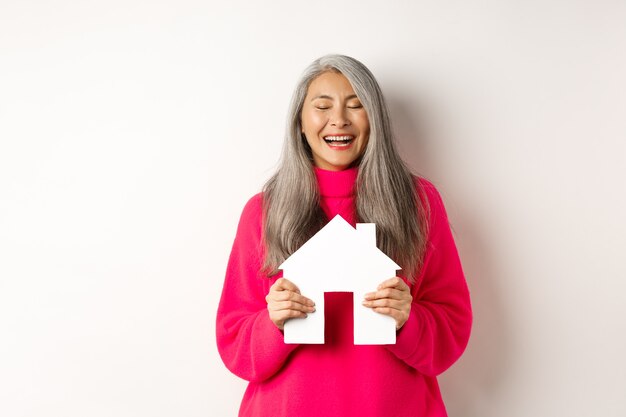 Real estate. Happy asian grandmother laughing with eyes closed, holding paper house model, dreaming about paper house model, standing over white background