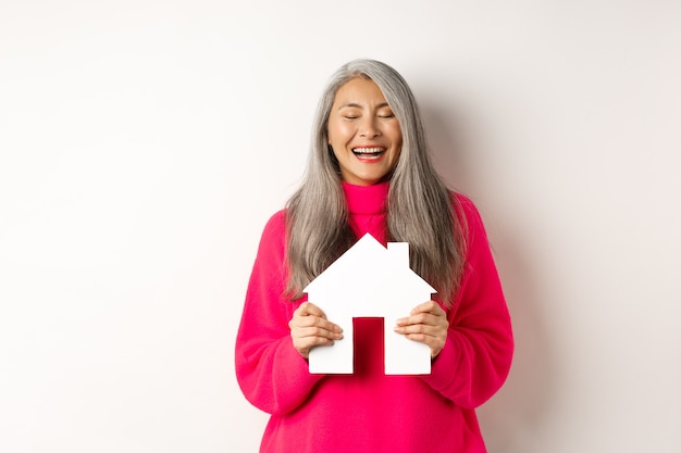 부동산. 행복한 아시아 할머니는 눈을 감고 웃고, 종이 집 모델을 들고, 종이 집 모델을 꿈꾸며, 흰색 배경 위에 서 있습니다.