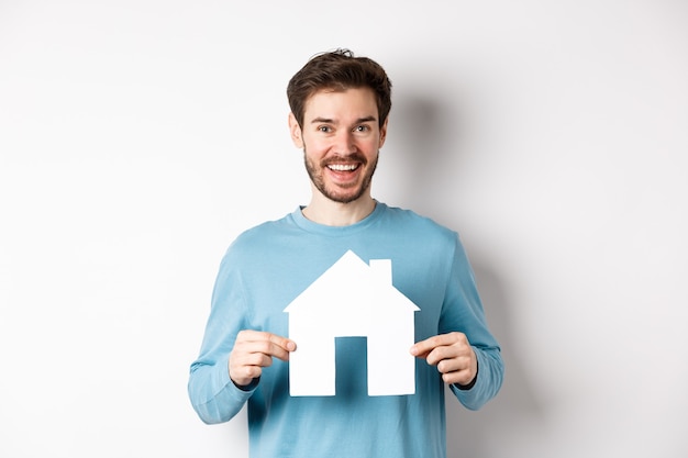 Концепция недвижимости и страхования. Красивый современный мужчина покупает недвижимость, улыбаясь и показывая вырез дома из бумаги, стоя на белом фоне.