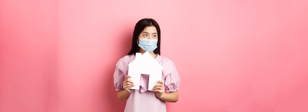 Free photo real estate and pandemic concept young asian woman in medical mask dreaming of home showing paper ho