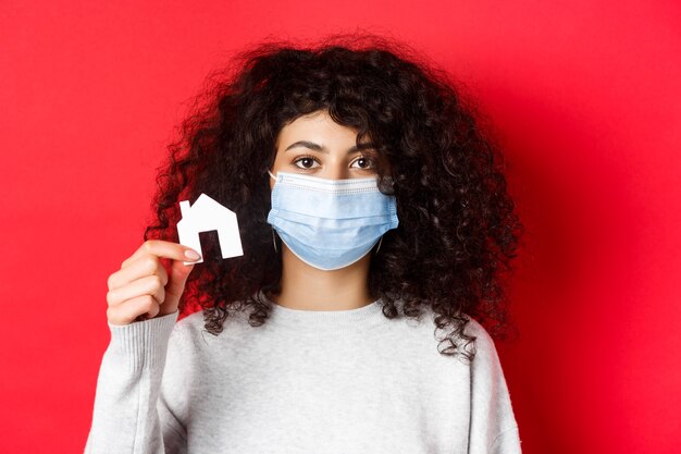 부동산 및 전염병 개념 의료 마스크를 쓴 젊은 여성이 작은 종이 집 컷아웃 스탠드를 보여줍니다.