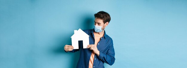 Real estate and quarantine concept young caucasian man in medical mask looking curious at paper hous