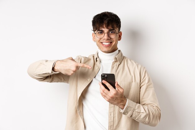 Real people. Handsome young man in glasses pointing finger at smartphone screen, showing online promo, standing on white background