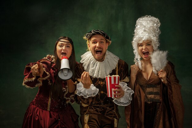 Real sport fans. Portrait of medieval young people in vintage clothing on dark background. Models as a duke and duchess, princess, royal persons. Concept of comparison of eras, modern, fashion.