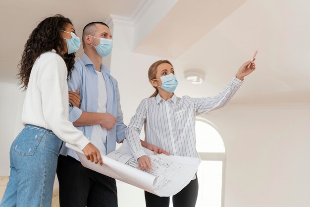 Realtor and couple with medical masks holding house plans