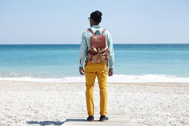 Снимок сзади стильного молодого афроамериканского туриста, стоящего на променаде на галечном пляже, смотрящего на огромный спокойный океан с чистой лазурной водой мирным утром, любуясь удивительным морским видом