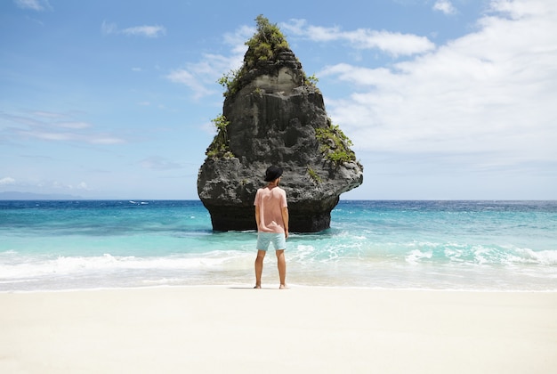 Free photo rear shot of stylish young man wearing trendy black hat standing barefooted on sandy beach in front of rocky cliff, waiting for unknown mysterious girl he met accidentally and fell in love with