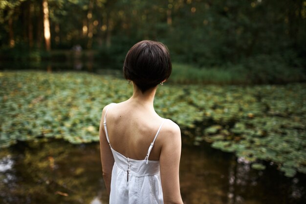 Rear shot of unrecognizable short haired young woman in strap white dress relaxing by pond in park, enjoying beautiful landscape and hot summer day. Back view of female walking outdoors alone