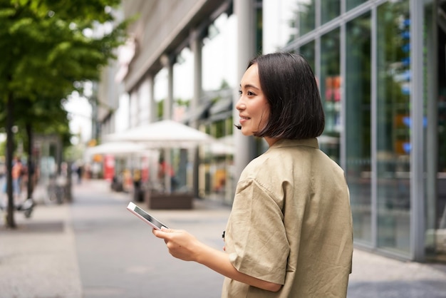 通りを下って街を歩いてスマートフォン ビュー f を保持している笑顔の若い女性のリア ショット