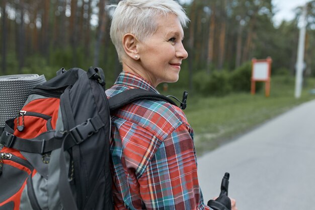 Rear view of adventurous middle aged woman with pixie haircut carrying backpack while hiking, going to spend weekends in mountains.