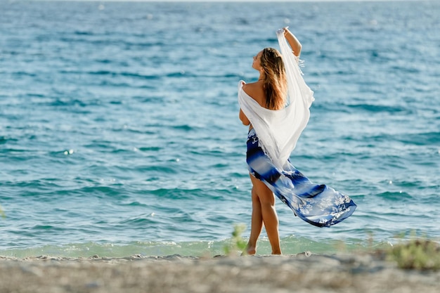 海のそばで夏の自由を楽しんでいるショールを持つ美しい女性の背面図