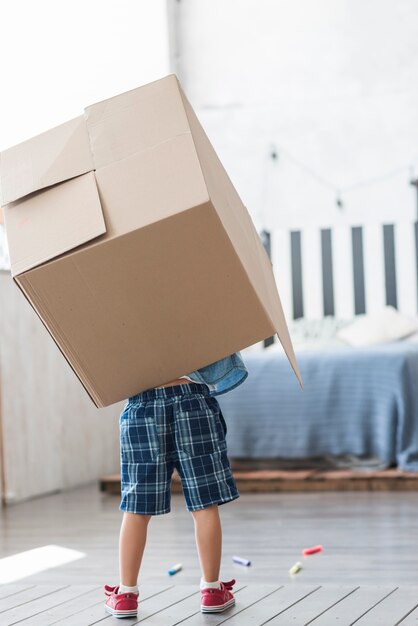 Вид сзади мальчик, держащий картонную коробку над спиной у себя дома