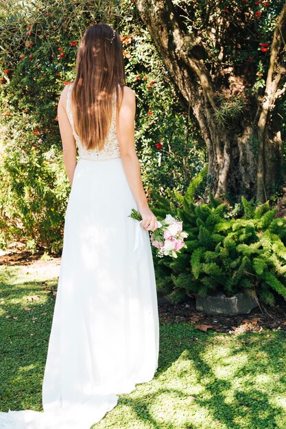 Rear view of a bride holding flower bouquet in hand standing in the park