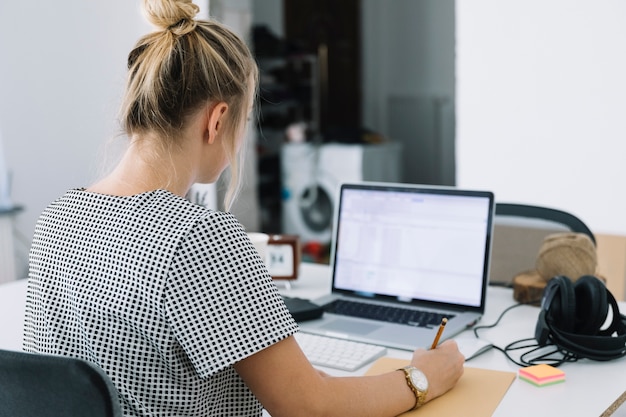 Foto gratuita la retrovisione di una donna di affari scrive le note sopra carta marrone con il computer portatile sullo scrittorio