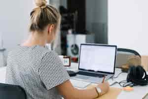 Foto gratuita la retrovisione di una donna di affari scrive le note sopra carta marrone con il computer portatile sullo scrittorio