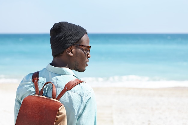 Rear view of carefree young dark-skinned traveler with leather backpack enjoying beautiful azure seascape while spending summer vacations by the sea, contemplating amazing view on sunny day