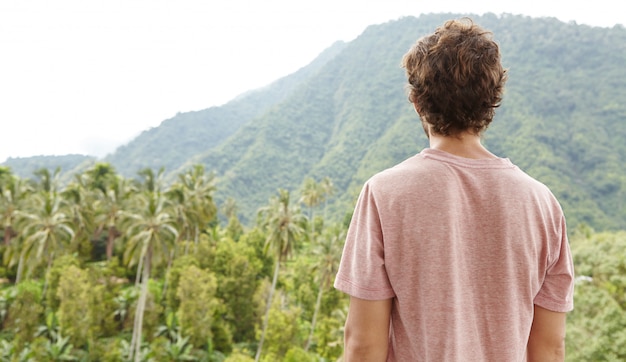 Rear view of Caucasian man in t-shirt standing outdoors in front of rainforest and contemplating beauties of exotic wild nature on sunny day. Tourist enjoying beautiful landscape during trekking trip