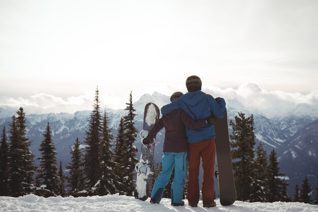 空に対して冬の間に山でスノーボードを保持しているカップルの背面図