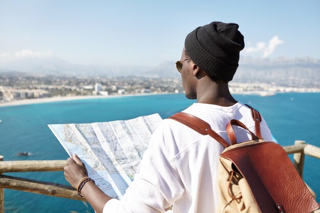 Rear view of of fashionable Afro American hiker with leather backpack on his shoulders holding paper guide, reading information about beautiful places and locations in front of him along sea coast