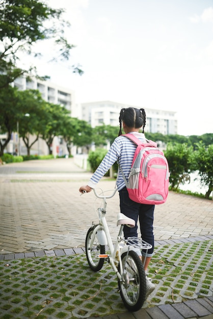 Rear view of female pupil wearing backpack and pulling bicycle in the street