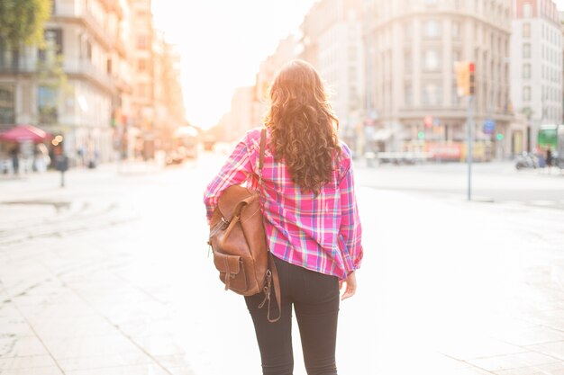 Rear view of female tourist with satchel walking over street and travelling alone. Curly-haired woman looking around and enjoying architecture of city. Travelling on weekend concept