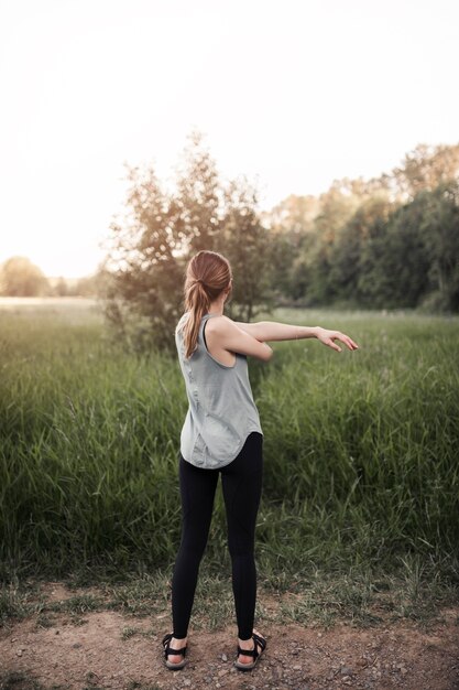 Вид сзади фитнес молодой женщины, растяжения ее руки, стоя в поле