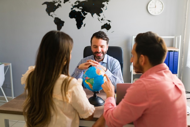 Free photo rear view of an hispanic young woman and man looking at countries on a globe while planning their trip abroad with a travel agent