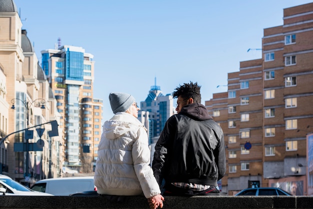 Free photo rear view of interracial young couple sitting on street in front of city buildings