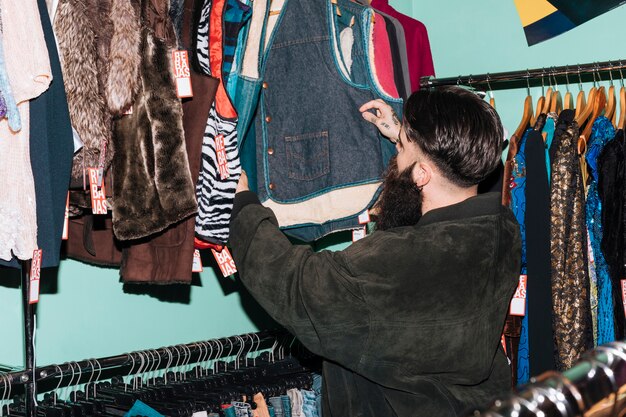 Вид сзади человека, выбирающего одежду, висящую на рельсе в магазине одежды