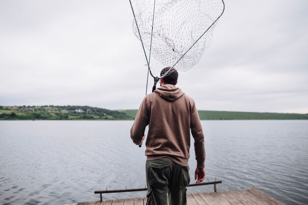 Вид сзади человека, держащего рыболовную сеть и удилище, глядя на озеро