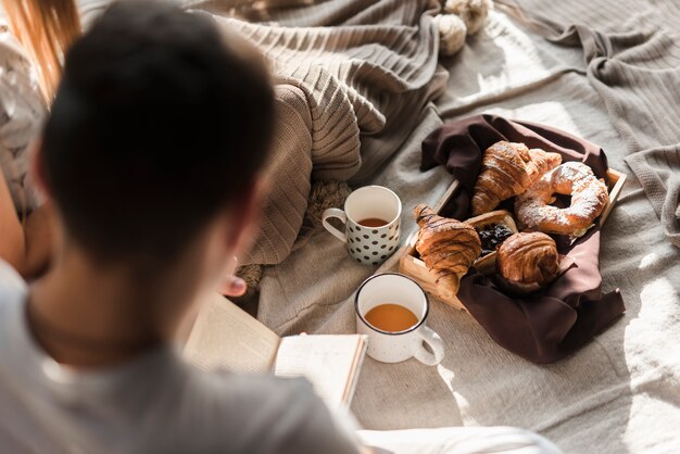 Rear view of a man reading book with breakfast on bed