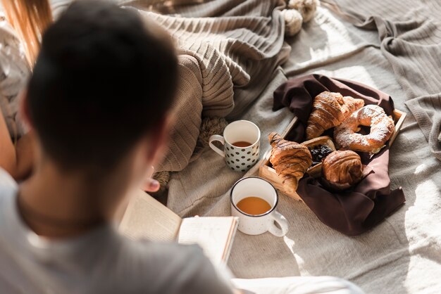 Вид сзади человек читает книгу с завтраком на кровати