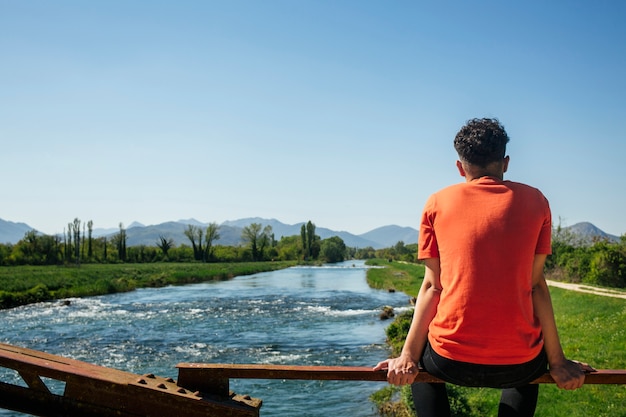 Вид сзади человека, сидящего на перилах возле идиллической реки