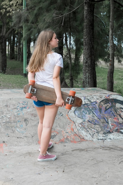 Rear view of modern girl holding skateboard standing in park