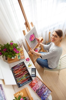 若い​女性​アーティスト​の​背面図​は​、​屋外​の​木製​イーゼル​に​座っている​間​、​彼女​の​スタジオ​で​乾いた​パステル​クレヨン​の​箱​から​ピンク​の​牡丹​の​スケッチ​を​描きます​。​創造性​と​趣味​の​コンセプト​。​ピンク​の​色合い​。