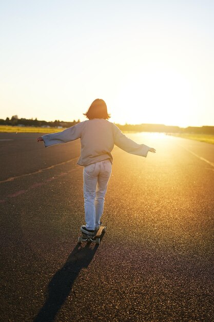 Фотография сзади молодой девушки, катающейся на скейтборде к солнечному свету счастливой молодой женщины на своем крейсере sk