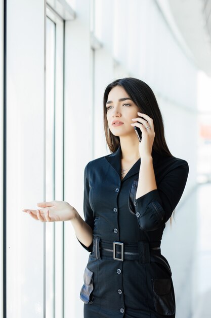 窓の外を見ながら、携帯電話を使用して話している若い労働者の背面図の肖像画。夕方、職場で忙しいビジネスコールの女性。