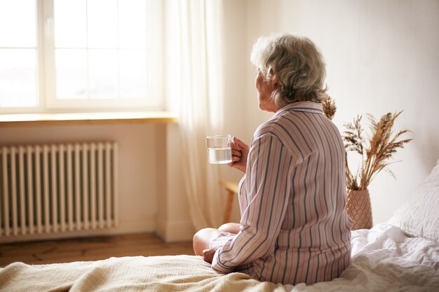 不眠症に苦しんで、睡眠薬を洗い流しているマグカップを保持している白髪のシニア60歳の女性の背面図。寝室に座って、水で薬を服用している高齢の引退した女性