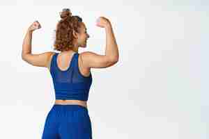 Free photo rear view of sports woman shows strong arms, flexing biceps in sportsbra and shorts, standing against white background.