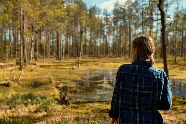 一人で屋外を歩いている、コートを着て松林でポーズをとって、沼の前に立って、春の日の美しい晴れた天気を楽しんでいるポニーテールの認識できない若い女性の背面図