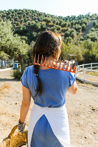 Rear view of woman holding rake on shoulder and basket in hand