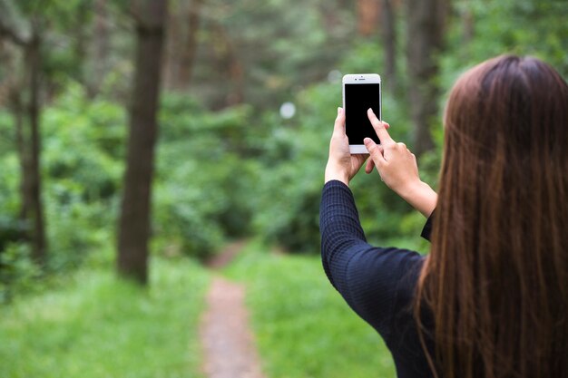 Вид сзади женщины, стоящей в лесу, касаясь экрана мобильного телефона
