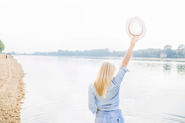 Rear view of woman standing near the lake raising hand holding hat