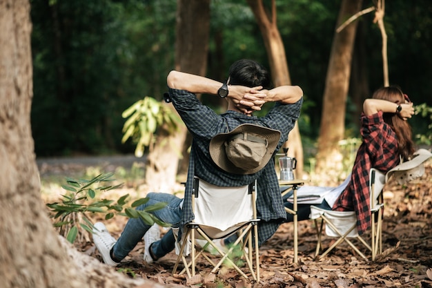 Free photo rear view, young asian teenager couple have relax time with camping trip, they are sitting and hands on back of neck on chair in front of backpack camping tent in forest