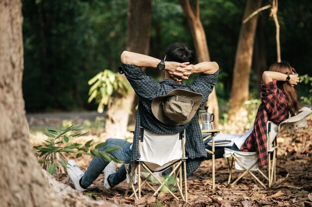 Rear view, Young Asian teenager couple have relax time with camping trip, They are sitting and hands on back of neck on chair in front of backpack camping tent in forest