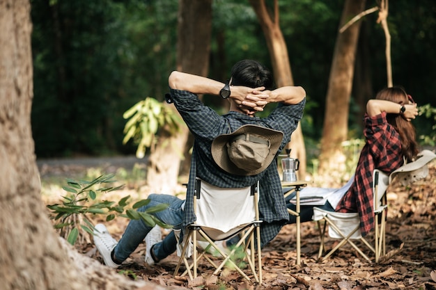 Вид сзади, молодая азиатская пара подростков отдыхает во время похода, они сидят, положив руки на затылок на стуле перед рюкзаком, палаткой в лесу