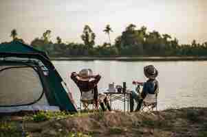 Free photo rear view of young backpacker couple sitting to relax at front of the tent near the lake with coffee set and making fresh coffee grinder while camping trip on summer vacation